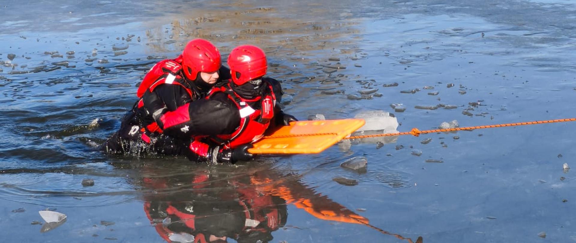 Ratownicy znajdują się w wodzie w rozbitym przeręblu. Jeden z nich trzyma poszkodowanego na pomarańczowej desce, która jest wciągana na lód za pomocą liny. W tafli lodu odbijają się sylwetki ratowników i ich czerwone kaski.