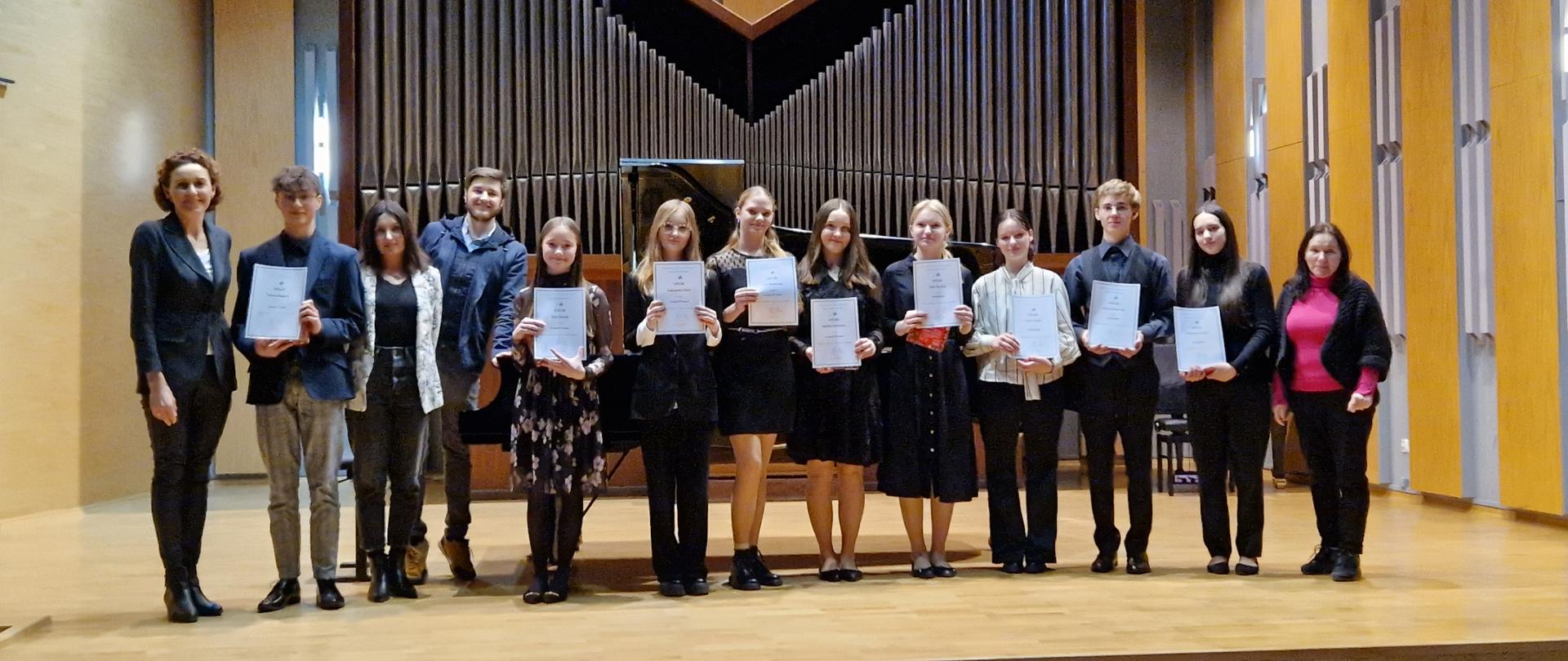 Zdjęcie grupowe. Uczestnicy konkursu wraz z pedagogami stoją w jednym rzędzie na scenie sali koncertowej, trzymając w swych dłoniach na wysokości piersi dyplomy. Na pierwszym planie puste dwa rzędy foteli. Za uczestnikami dwa fortepiany i organy w tle