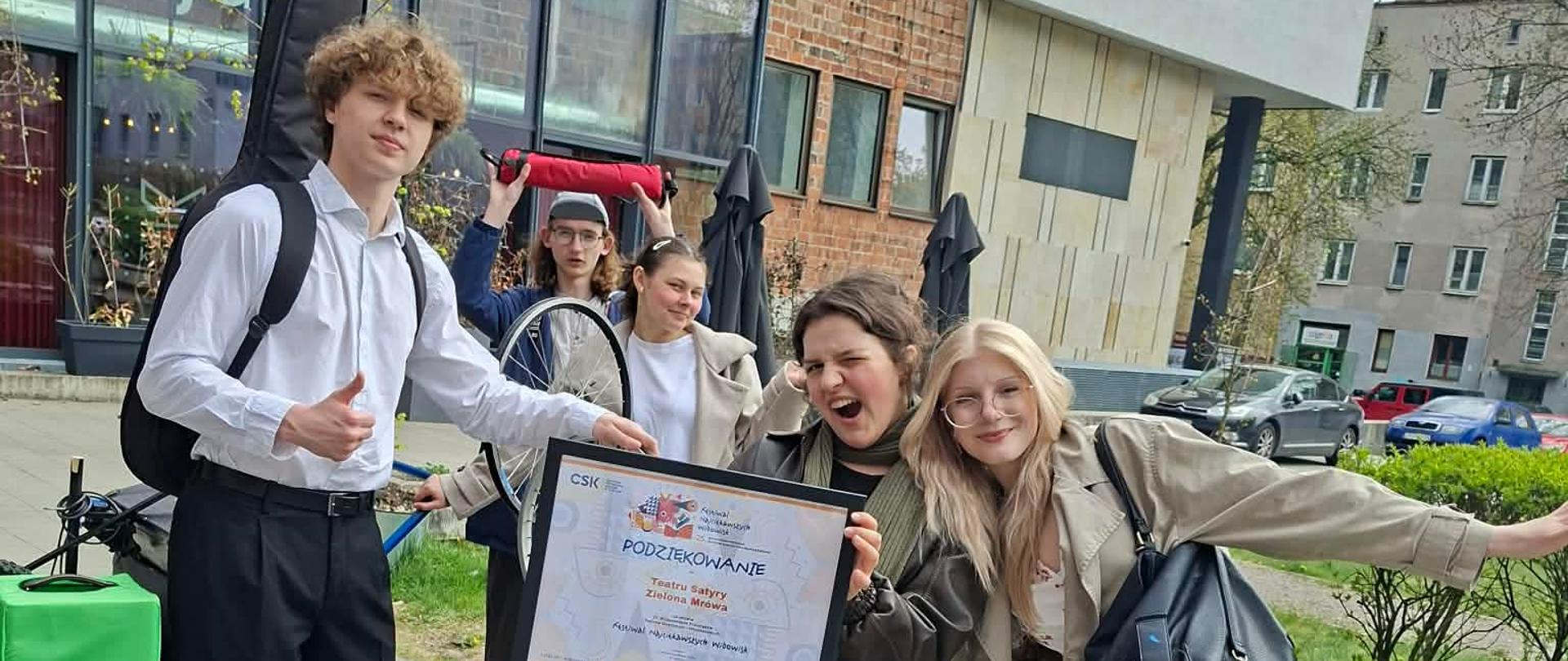 Na tle budynku i zieleni do zdjęcia pozują robiąc wesołe miny trzy dziewczyny i dwóch chłopców z lewej strony . W rękach trzymają czarna ramkę z podziękowaniem