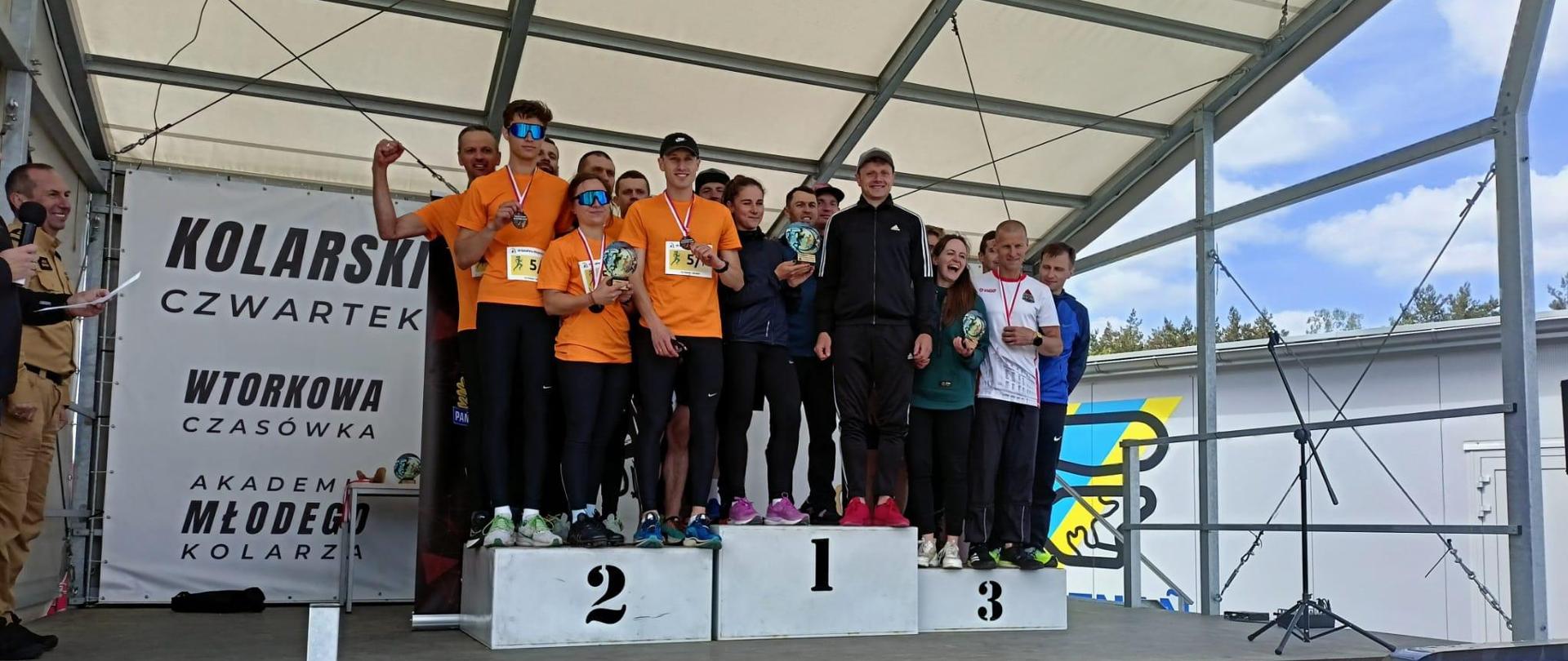 Pod zadaszeniem na podium stoją zawodnicy w ubraniach sportowych, z trzech pierwszych drużyn w biegu sztafetowym.