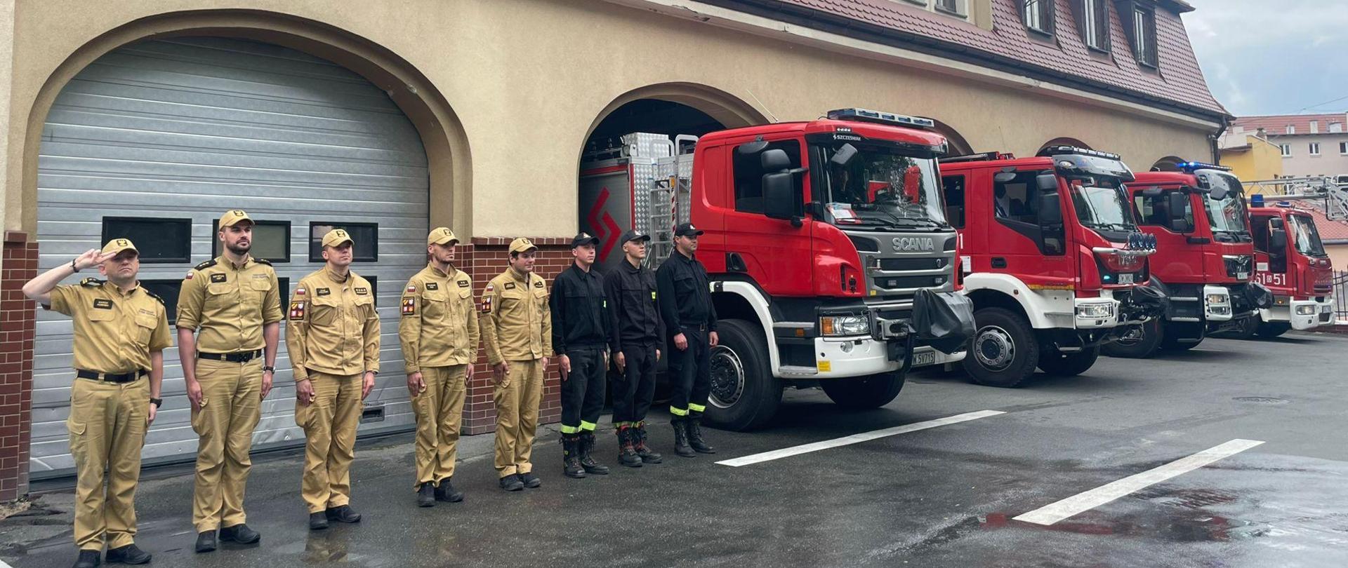 Plac przed budynkiem Komendy. Widok na bramy garażowe. Od lewej strony w rzędzie stoi ośmiu strażaków. Za nimi stoją cztery samochody pożarnicze.