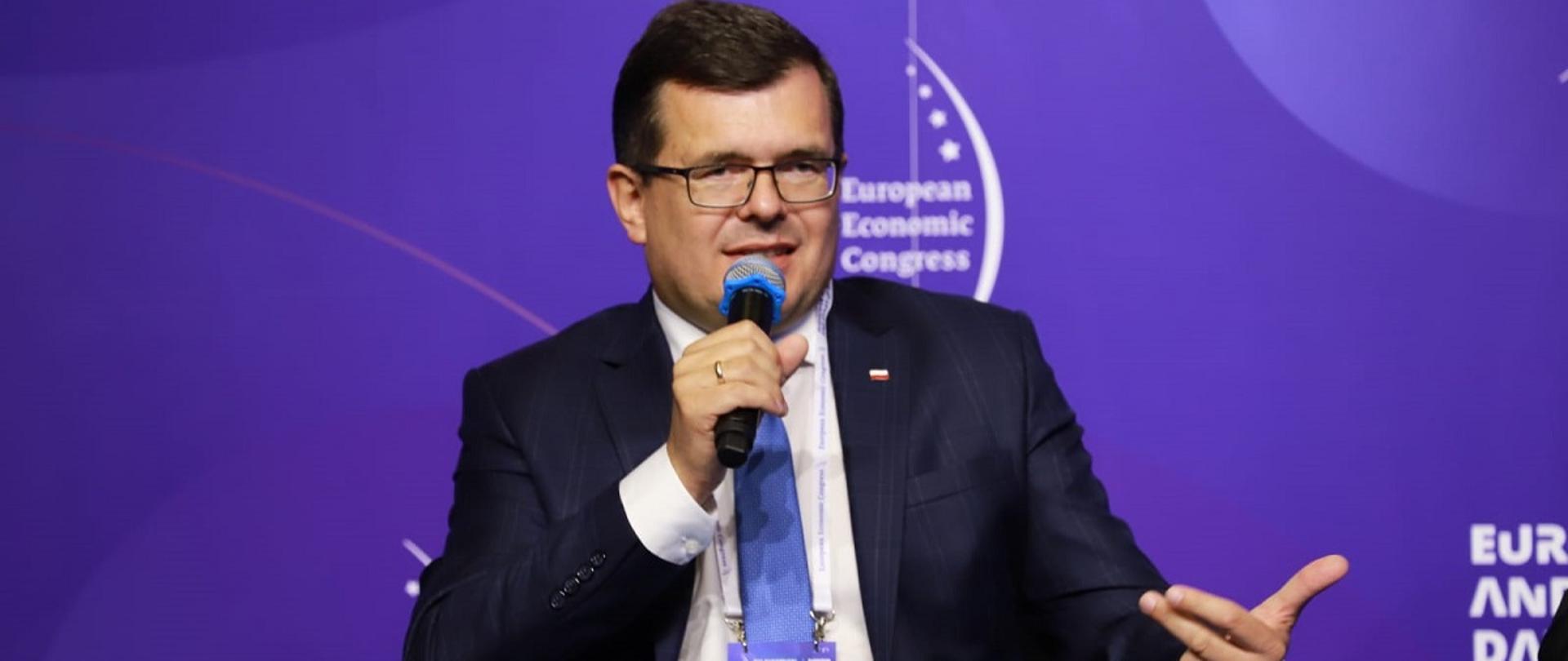 Wiceminister rozwoju i technologii Piotr Uściński podczas dyskusji poświęconej budownictwu na Europejskim Kongresie Gospodarczym w Katowicach, wiceminister siedzi i trzyma w dłoni mikrofon, za jego plecami baner z logo EKG
