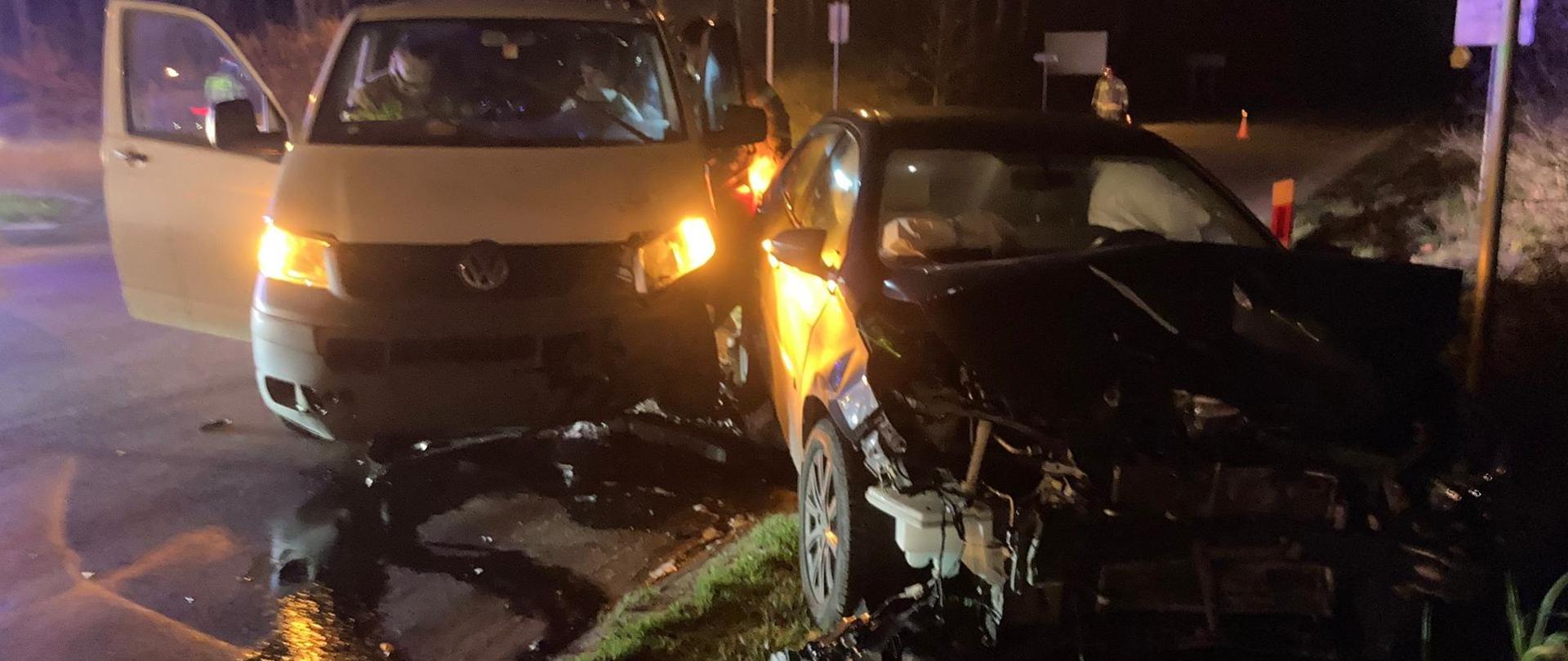 Na zdjęciu panuje noc. Na pierwszym planie widoczne są dwa rozbite pojazdy czarny samochód osobowy i biały bus. Stoją na jednym pasie drogowym. W tle widoczni strażacy ubrani w ubrania specjalne koloru piaskowego, zabezpieczają przy pomocy pachołków miejsce zdarzenia.