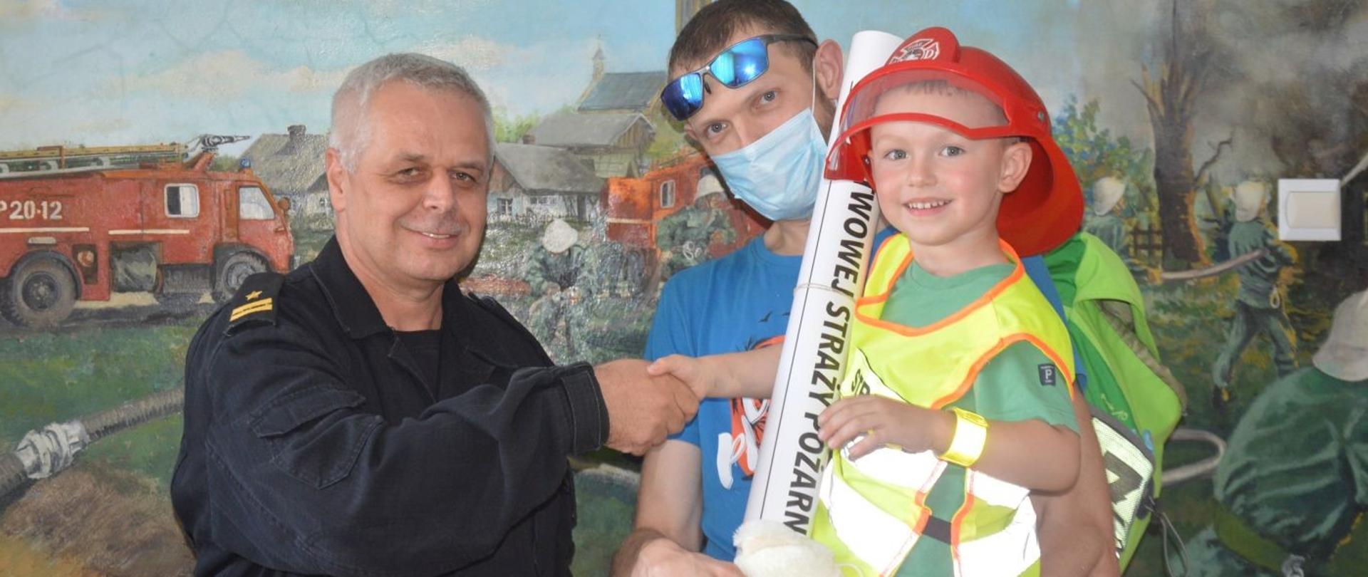 Wspieramy leczenie Gosi. Zastępca komendanta powiatowego PSP w swoim biurze ściska dłoń 3-letniemu synowi laureata licytacji "Dnia ze strażakami w Komendzie Powiatowej PSP w Rawiczu". Chłopiec jest na rękach swojego taty, ubrany jest w czerwony hełm i żółtą odblaskową kamizelkę. Tata ma niebieska koszulkę z krótkim rękawkiem a usta i nos ma zasłonięte maseczką. Wszyscy stoją na tle obrazu ściennego, który przedstawia akcję ratowniczo-gaśniczą.