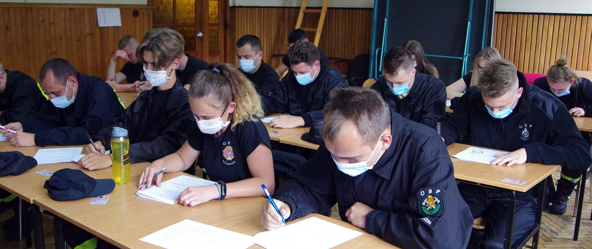 Widok z boku. Członkowie OSP w ubraniach koszarowych i maseczkach ochronnych siedzą w ławkach na sali szkoleniowej i rozwiązują test wiedzy.
