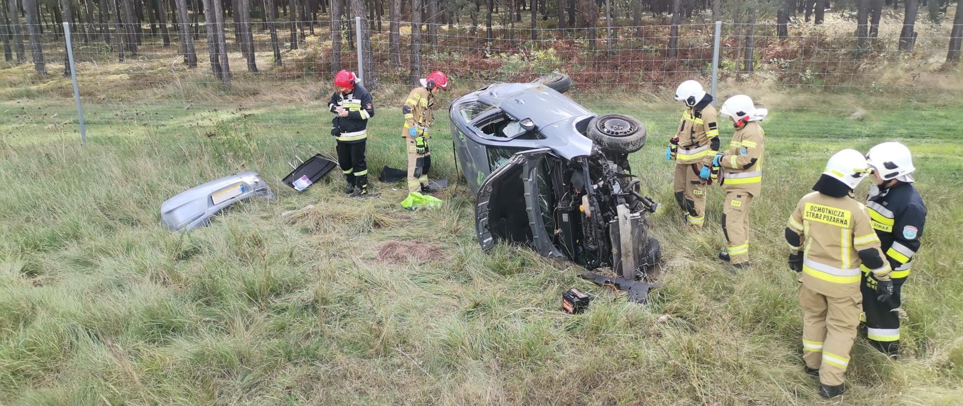 Zdjęcie przedstawia samochód osobowy leżący na boku na poboczu drogi ekspresowej. Wokół niego stoi sześciu strażaków wykonujących czynności.