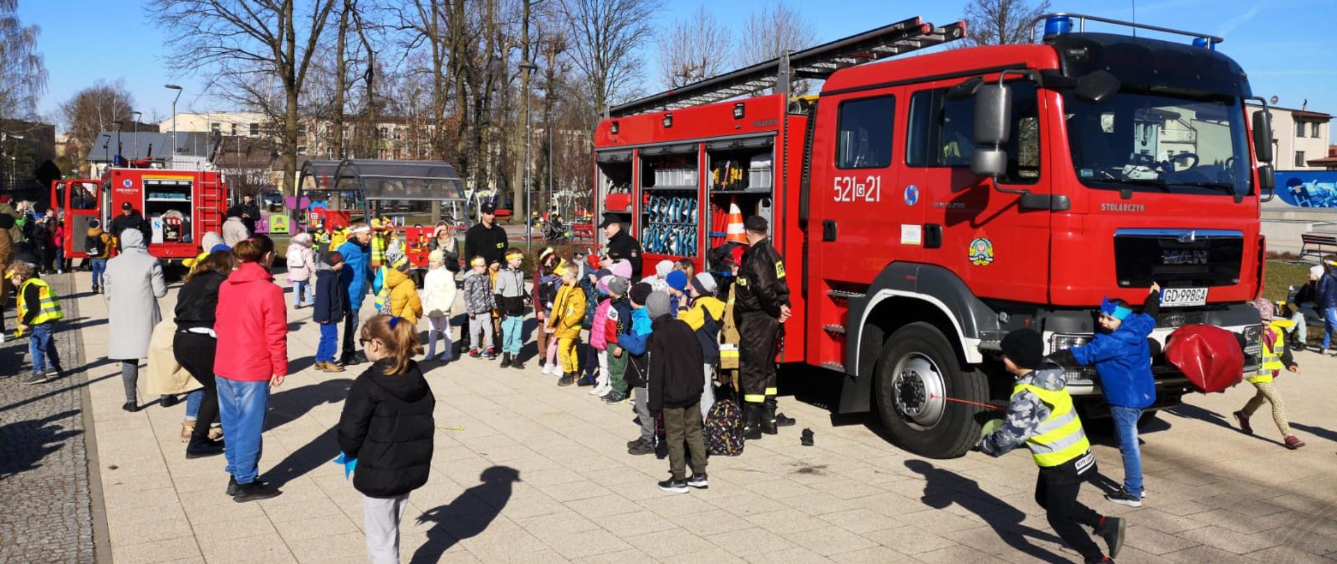 Zdjęcie przedstawia grupę dzieci z opiekunami podczas festynu. W centrum zdjęcia samochód pożarniczy, strażacy opowiadają dzieciom o pracy, w tle samochód pożarniczy z otwartymi skrytkami.