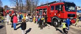 Zdjęcie przedstawia grupę dzieci z opiekunami podczas festynu. W centrum zdjęcia samochód pożarniczy, strażacy opowiadają dzieciom o pracy, w tle samochód pożarniczy z otwartymi skrytkami.