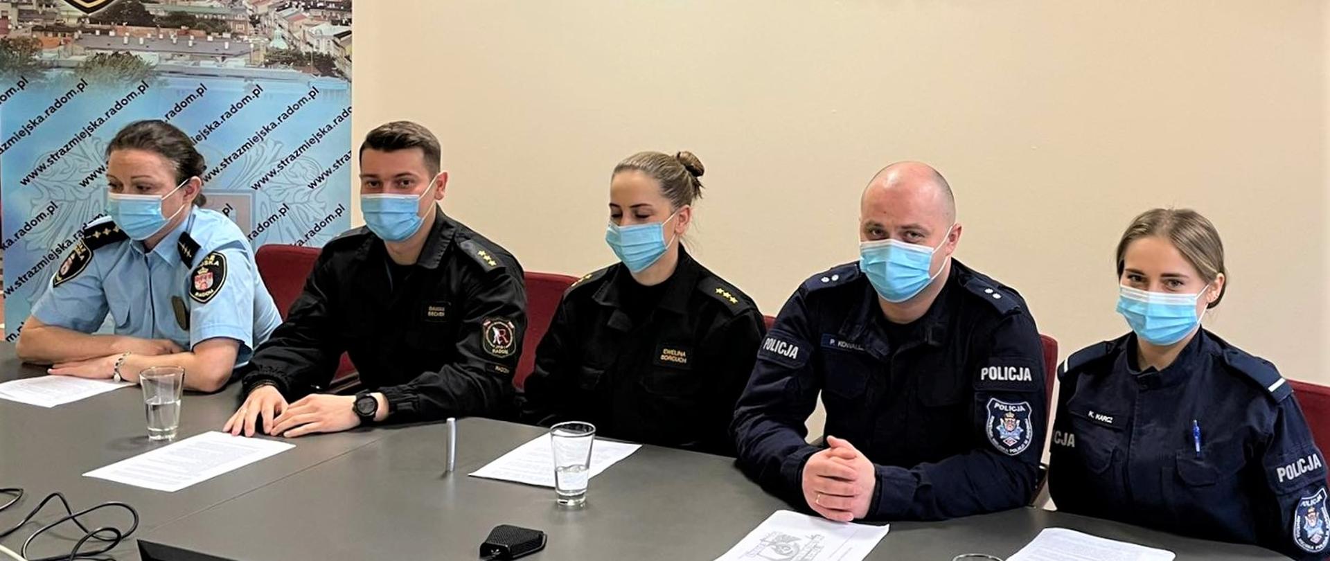 funkcjonariusze siedzą przy stole, od lewej Straż Miejska, Państwowa Straż Pożarna, Policja. Przed nimi leżą kartki z notatkami. Wszyscy mają założone maseczki ochronne.

