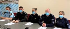 funkcjonariusze siedzą przy stole, od lewej Straż Miejska, Państwowa Straż Pożarna, Policja. Przed nimi leżą kartki z notatkami. Wszyscy mają założone maseczki ochronne.
