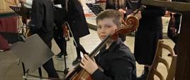 Uczeń Jan Jasiński trzymający wiolonczelę, w tle grupa kilku osób z szkolnej orkiestry kameralnej