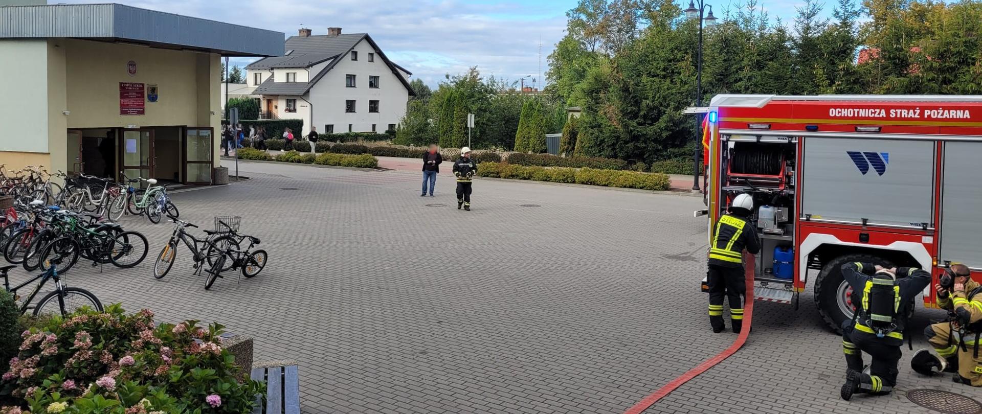 Plac przed szkołą wyłożony kostką brukową, z lewej strony widać tył czerwonego samochodu straży pożarnej z włączoną niebieską sygnalizacją świetlną. Przy samochodzie stoi strażak obsługujący pojazd ubrany w czarne ubranie specjalne z żółtymi pasami i białym hełmie na głowie, obok niego klęczy dwóch strażaków ubierających sprzęt ochrony układu oddechowego. Jeden ubrany w czarne ubranie specjalne koloru czarnego z żółtymi pasami, drugi w ubraniu specjalnym koloru piaskowego. Po lewej stronie widać zaparkowane rowery oraz otwarte drzwi wejściowe do budynku szkoły nad drzwiami urzędowe tablice informacyjne oraz godło państwowe, budynek szkoły w kolorach beżowym oraz zielonym. Na środku placu widać strażaka ubranego w czarne ubranie specjalne w żółte pasy na głowie biały hełm, z nim stoi osoba cywilna w oddali widać budynek mieszkalny a na jego tle uczniów idących chodnikiem. 