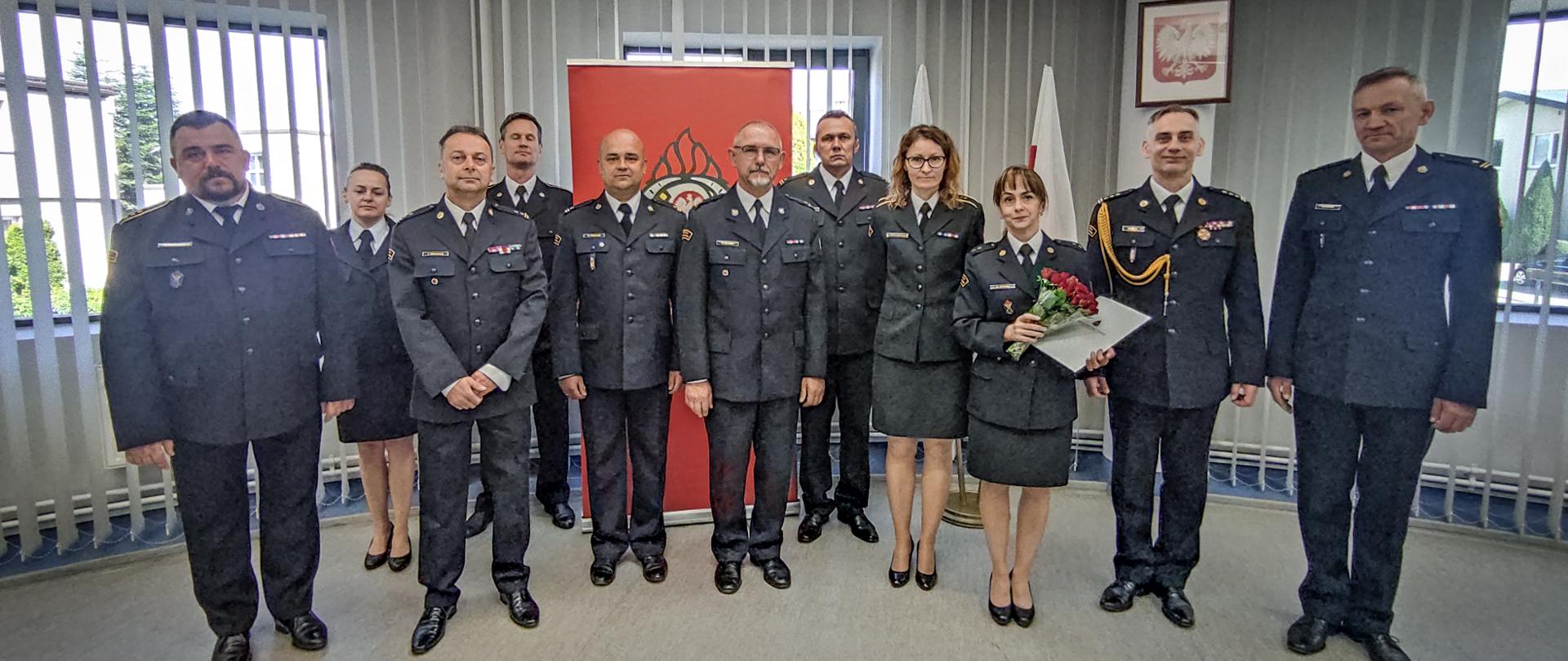 Zdjęcie zrobione w pomieszczeniu strażnicy na tle godła Rzeczpospolitej Polskiej, baneru Komendy Powiatowej PSP w Lublińcu oraz dwóch Flag Państwowych. Na pierwszym planie dziesięciu funkcjonariuszy w umundurowaniu wyjściowym i jeden w umundurowaniu galowym.