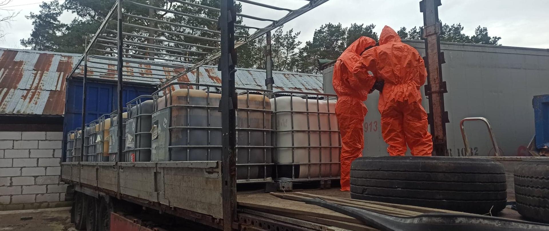 Zdjęcie przedstawia naczepę samochodu ciężarowego bez plandeki zaparkowaną na betonowym placu. W wnętrzu naczepy znajdują się 16 zbiorników typu Mauzer każdy o pojemności 1000 litrów. Wewnątrz nich znajduje się nieznana substancja. Zbiorniki są ustawione w dwóch rzędach jeden obok drugiego. Przed zbiornikami znajduje się dwóch ratowników ubranych w pomarańczowe kombinezony przeciwchemiczne, którzy badają nieznaną substancję w zbiornikach. Na podłodze przed ratownikami widoczne są także dwie opony samochodu ciężarowego. Za naczepą znajdują się zabudowania pokryte szarą blachą, częściowo zardzewiałe. Obok postawiony jest kontener ciężarowy. 