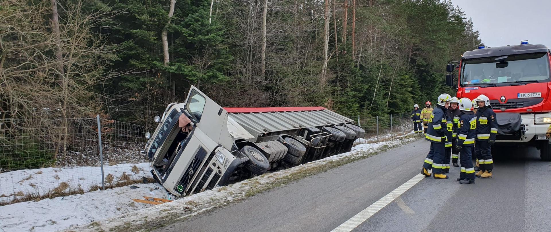 Zdjęcie przedstawia drogę ekspresową S7. Po lewej stronie, w rowie leży na boku uszkodzony samochód ciężarowy z naczepą. Po prawej, na jezdni stoi grupa strażaków, za którymi widać przód samochodu pożarniczego.