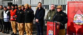 Wzdłuż ogrodzenia stoją w dwóch szeregach mężczyźni. Za mównicą stoi Komendant Miejski. Strażacy ubrani są w piaskowe mundury z czarnym polarem na głowach mają czapki z daszkiem. Obok Komendant stoi prezes Zakładów Mechanicznych ubrany w zieloną kurtkę oraz prezydent miasta. Z tyłu bilbord Państwowej Straży Pożarnej oraz budynki zakładów.