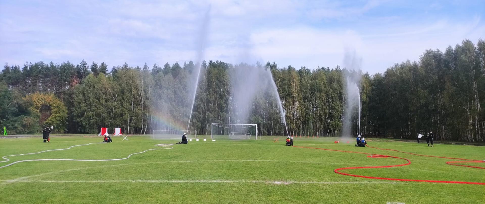 Na boisku piłkarskim widać rozwinięte węże strażackie, w oddali na końcu węży widać sylwetki strażaków trzymających węże z których wypływają trzy strumieni wody w górę.