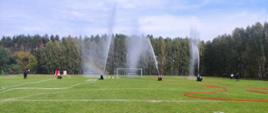 Na boisku piłkarskim widać rozwinięte węże strażackie, w oddali na końcu węży widać sylwetki strażaków trzymających węże z których wypływają trzy strumieni wody w górę.