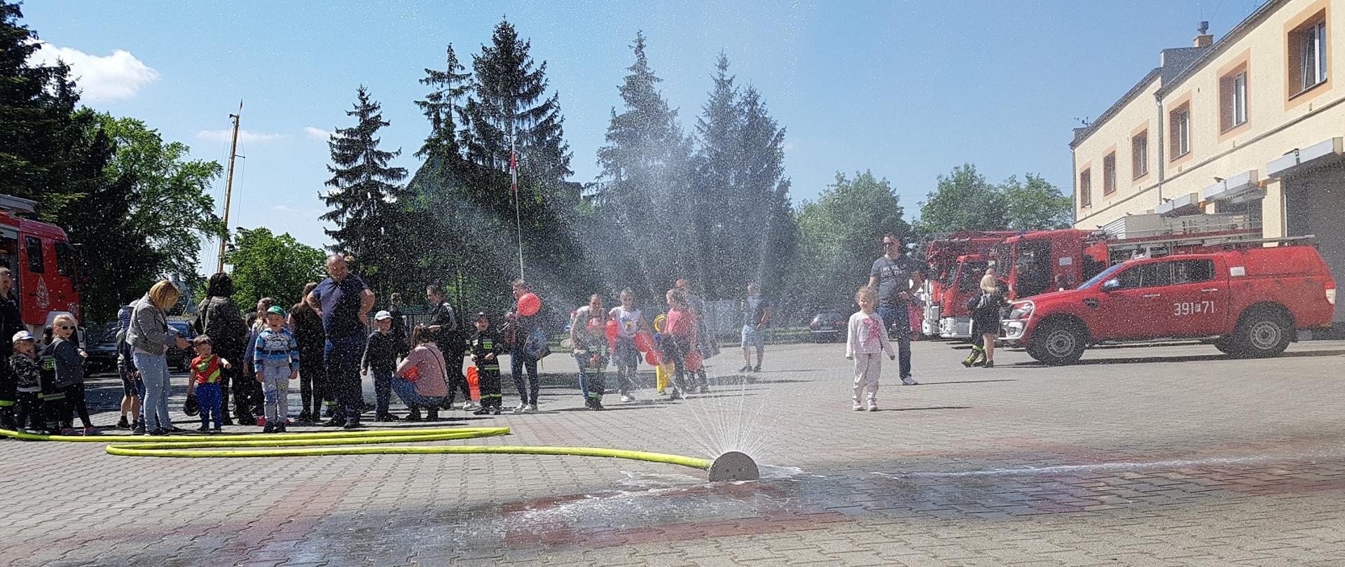 Plac wewnętrzny KP PSP w Słubicach z widoczną rozstawioną kurtyną wodną. Z tyłu samochody strażackie i w tle dzieci z opiekunami.