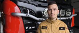 Starszy aspirant stoi na tle samochodu strażackiego, ubrany jest w piaskowy mundur na którym widać dystynkcje oraz imiennik