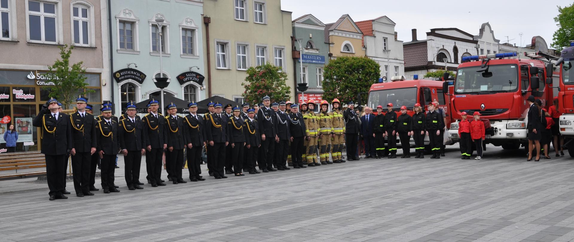 Zdjęcie przedstawia strażaków ubranych w mundury wyjściowe oraz mundury specjalne. Strażacy stoją na Starym Rynku w Pucku. Ratownicy stoją ustawieni w dwuszeregu na tle budynków oraz samochodów strażackich.