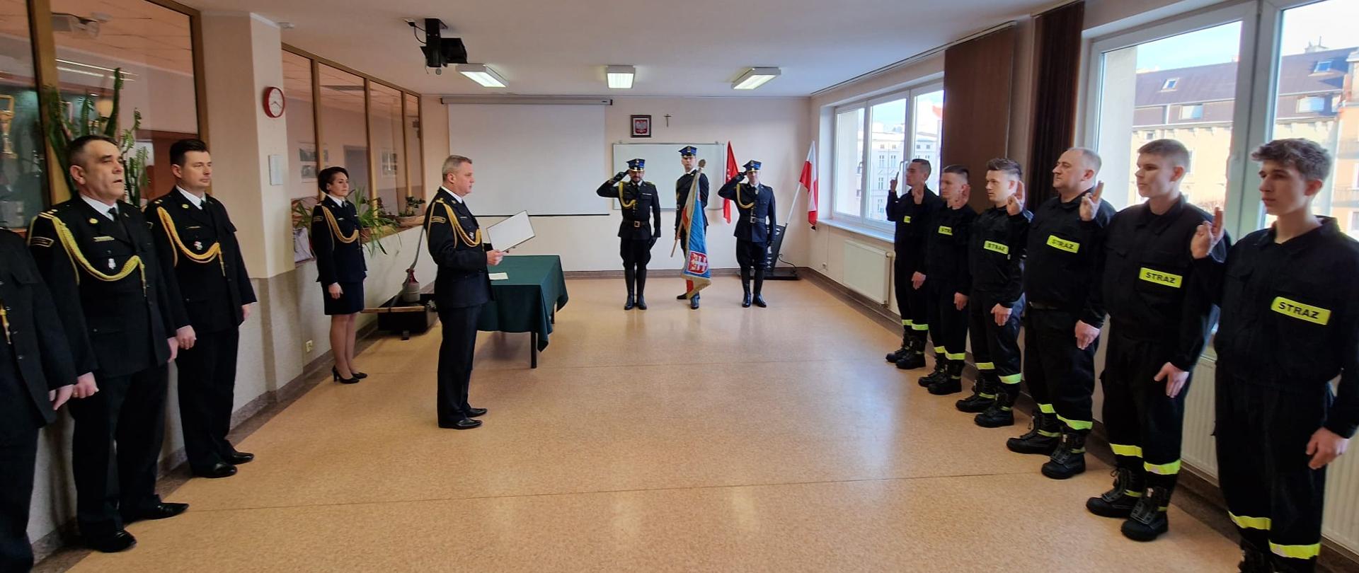 Komendant Miejski odczytuje akt ślubowania na świetlicy JRG sześciu funkcjonariuszom. W tle poczet sztandarowy.