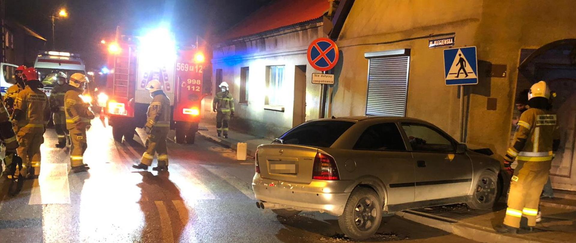 Zdjęcie przedstawia zdarzenie drogowe wieczorem, w wyniku którego samochód osobowy marki Opel wjechał na chodnik uderzając w znak drogowy i budynek. Strażacy zabezpieczają miejsce zdarzenia oraz uszkodzony samochód. 