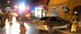 Zdjęcie przedstawia zdarzenie drogowe wieczorem, w wyniku którego samochód osobowy marki Opel wjechał na chodnik uderzając w znak drogowy i budynek. Strażacy zabezpieczają miejsce zdarzenia oraz uszkodzony samochód. 