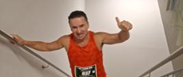 Uczestnik zawodów sportowych biegnie po schodach podczas konkurencji wymagającej pokonania klatki schodowej. Ma na sobie pomarańczowy strój sportowy z numerem startowym oraz obuwie biegowe. Zawodnik trzyma się poręczy i unosi rękę w geście zachęty podczas pokonywania kolejnych stopni.