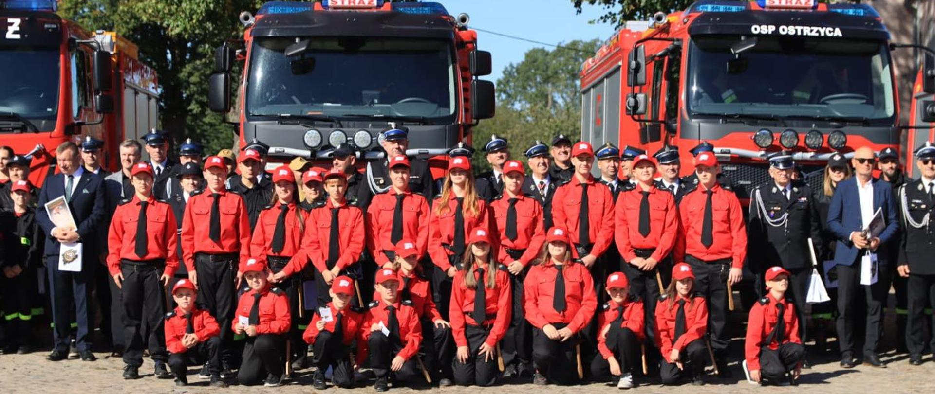 Na zdjęciu widać dużą grupę strażaków i młodzieży pożarniczej pozujących wspólnie do pamiątkowego zdjęcia. W pierwszym rzędzie klęczą i stoją młodzi ludzie w czerwonych koszulach, czarnych spodniach, czarnych krawatach i czerwonych czapkach z białym symbolem. Za nimi stoją strażacy w ciemnych, galowych mundurach z czapkami z daszkiem. W tle widoczne są trzy duże, czerwone wozy strażackie z napisem „STRAŻ”, a na jednym z nich dodatkowo „OSP Ostrzyca”. Cała scena odbywa się na brukowanym placu w słoneczny dzień, w otoczeniu drzew i budynków.