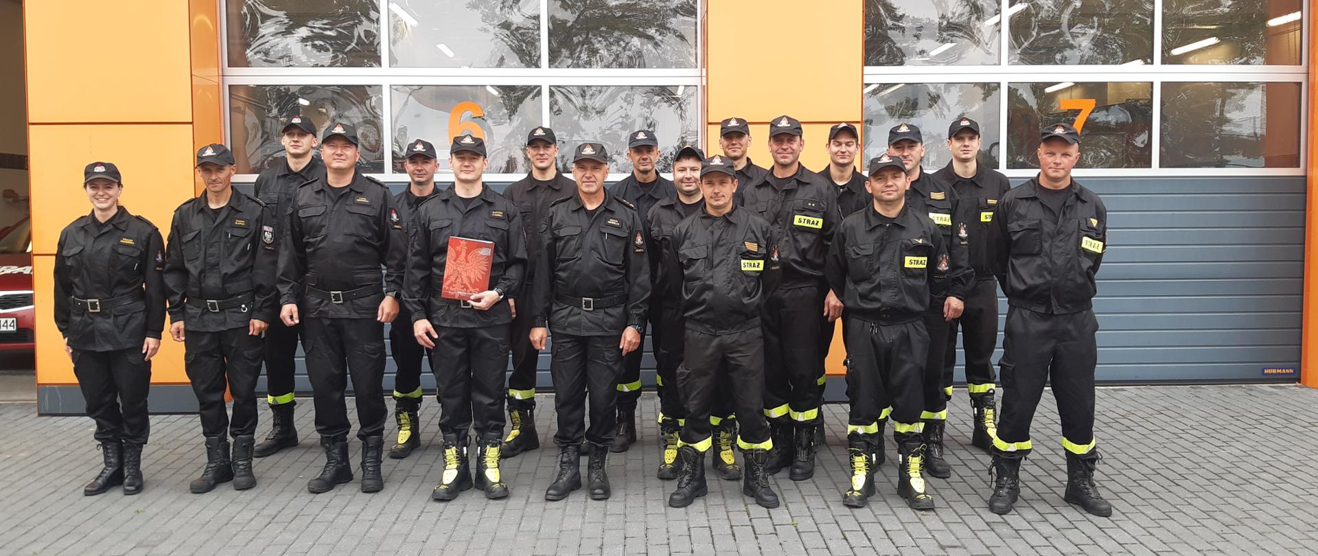 Na zdjęciu funkcjonariusze KP PSP w Wolsztynie, ubrani w czarne mundury. Stoją na wprost, zdjęcie grupowe. W tle bramy garażowe jednostki.