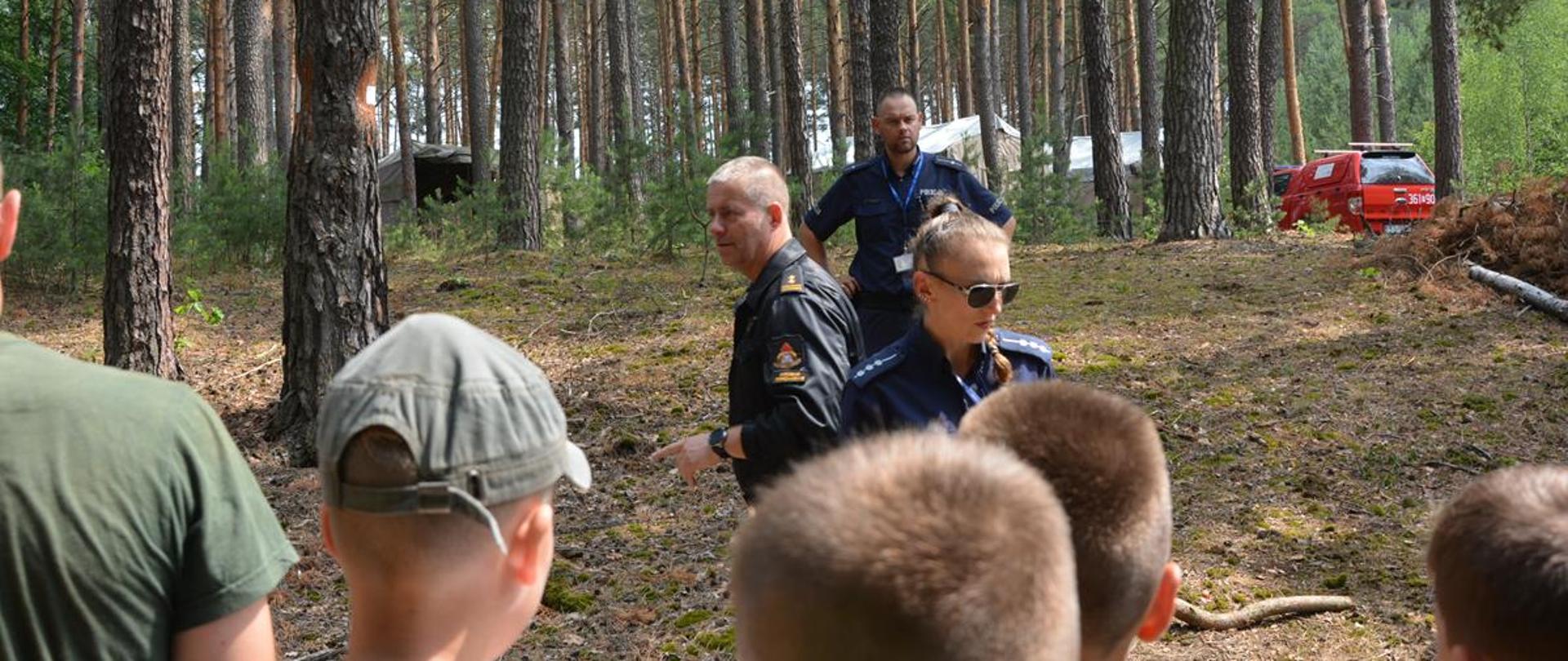 Zdjęcie przedstawia funkcjonariusza Komendy Powiatowej Państwowej Straży Pożarnej w Krośnie Odrzańskim, funkcjonariuszy Komendy Powiatowej Policji w Krośnie Odrzańskim oraz harcerzy podczas kontroli w ramach akcji „Bezpieczne Wakacje 2022”. Zdjęcie wykonane zostało na tle lasu.