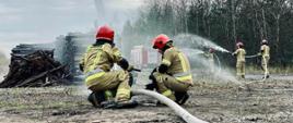 Na pierwszym planie widocznych dwóch kucających strażaków podających gaśniczy prąd wody z działka na składowisko odpadów. W tle widoczne dodatkowe siły i środki podające dwa prądy gaśnicze wody na tle lasu.