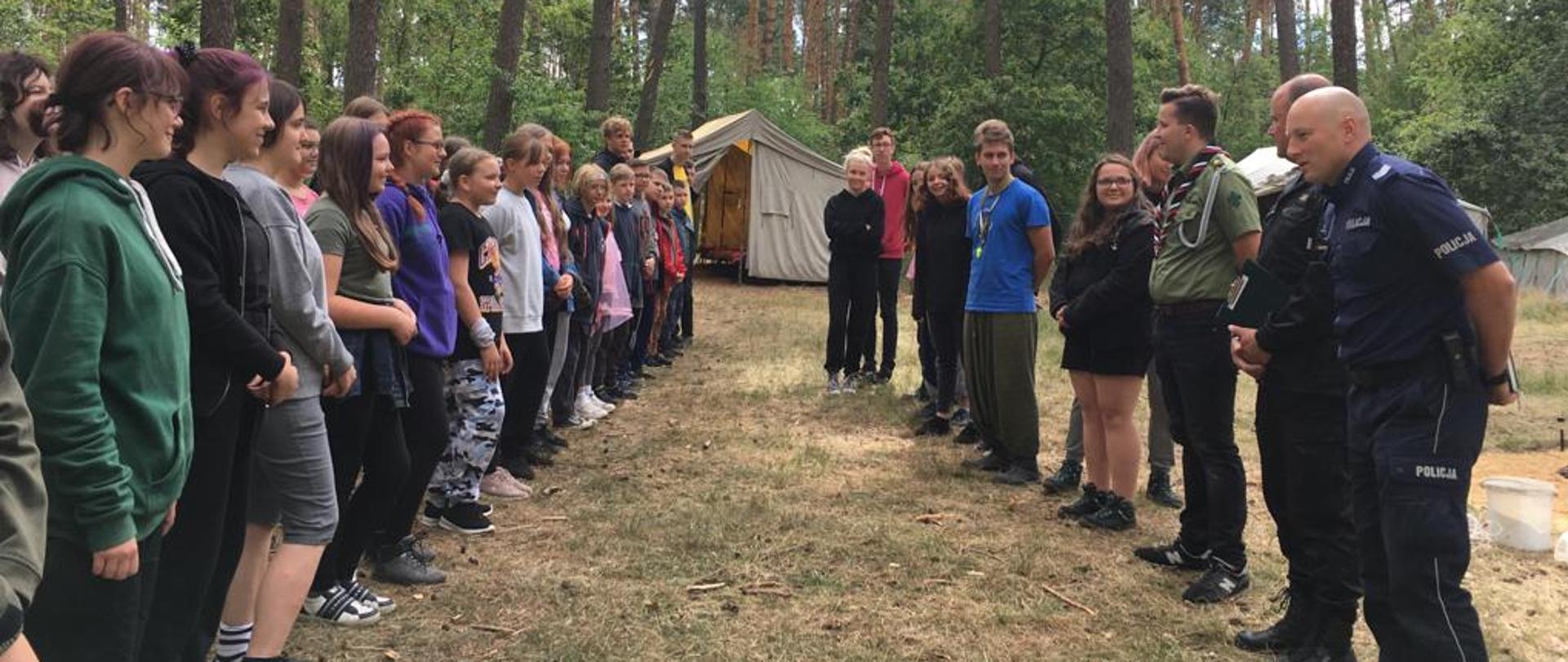 Na zdjęciu wizytacja obozu harcerskiego przez służby mundurowe. Strażak i policjant przekazuje informacje o zagrożeniach na obozowisku, harcerze stoją w lesie w ubraniach cywilnych po przeciwnej stronie służby mundurowe w tle widać namiot.