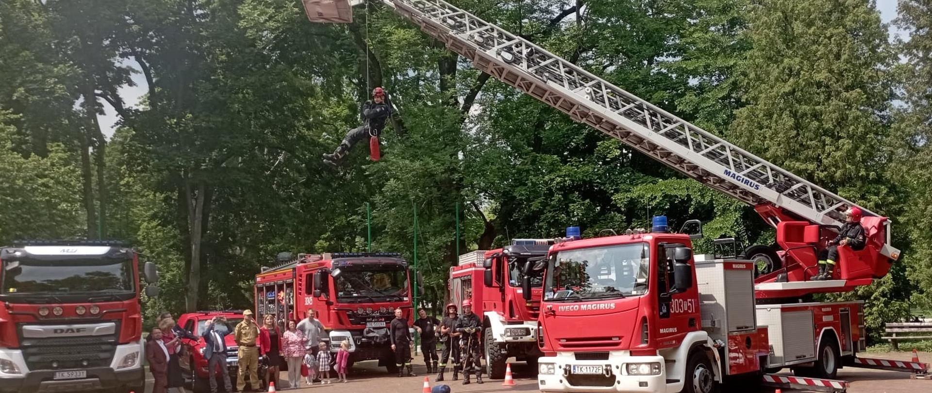 Na zdjęciu widać drabinę mechaniczna rozłożoną, na której podwieszony jest ratownik pozorujący osobę poszkodowaną. Za nią stoją obserwatorzy pokazu oraz cztery samochody pożarnicze. 