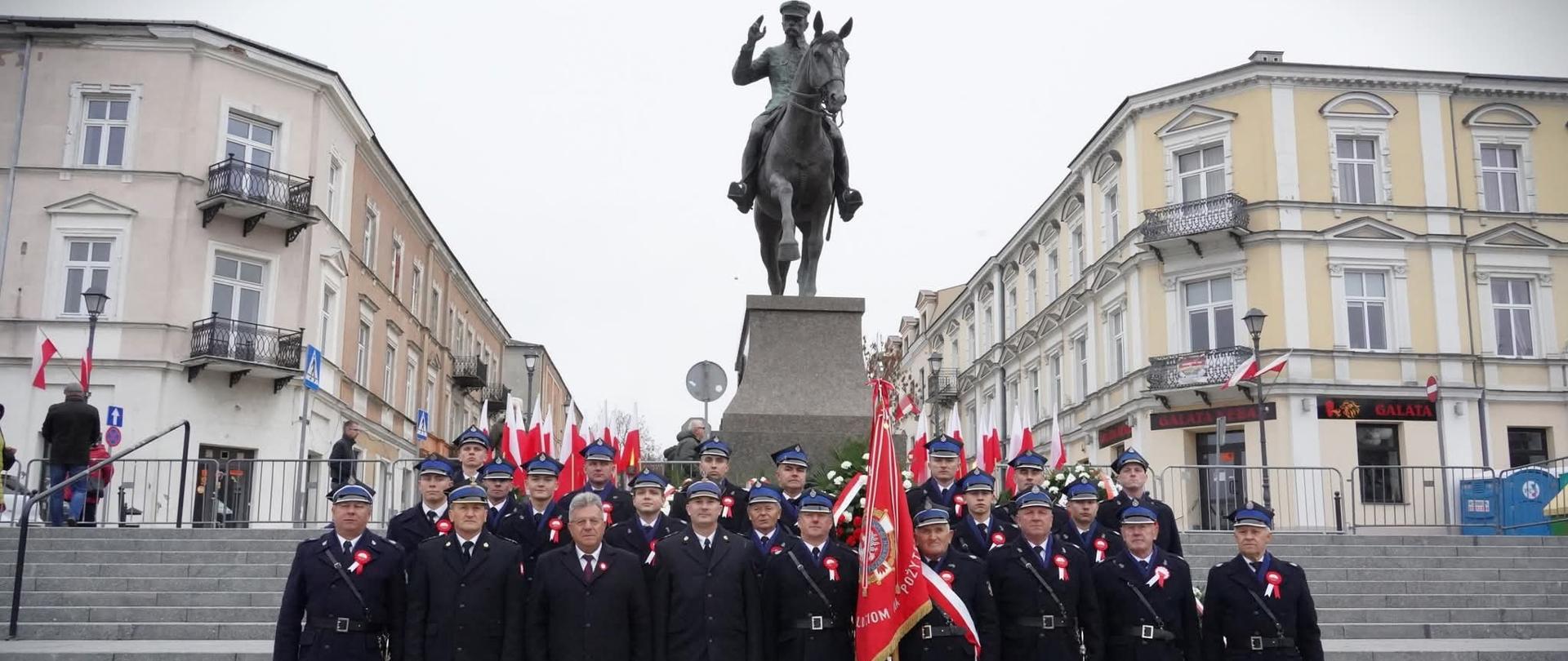 Strażacy biorący udział w uroczystościach pozują pod pomnikiem marszałka Józefa Piłsudskiego