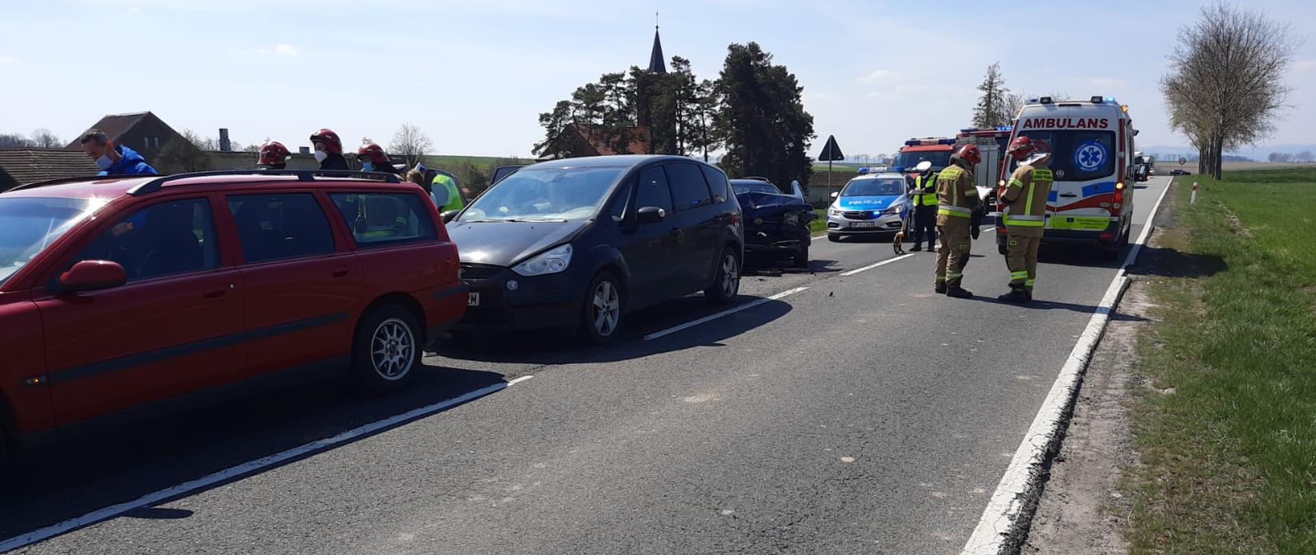 Zdjęcie przedstawia miejsce zdarzenia po wypadku samochodów osobowych. W głębi zdjęcia widać funkcjonariuszy straży pożarnej, ambulans pogotowia ratunkowego, samochód policyjny oraz samochód straży pożarnej.
