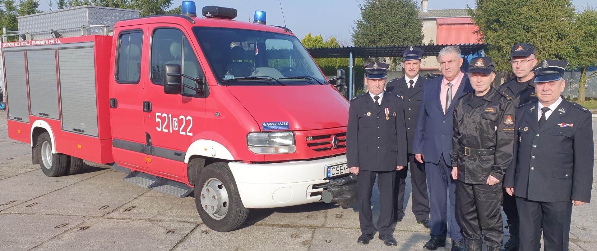Zdjęcie wykonano za dnia na placu Komendy Powiatowej Państwowej Straży Pożarnej. Przed lekkim samochodem pożarniczym stoją Komendanci Komendy Powiatowej Państwowej Straży Pożarnej w Sępólnie Krajeńskim oraz strażacy z Ochotniczej Straży Pożarnej ubrani w mundury wyjściowe.