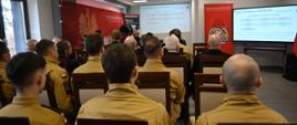 Strażacy i zgromadzeni goście siedzą na sali konferencyjnej. Patrzą na przedstawianą prezentację. 