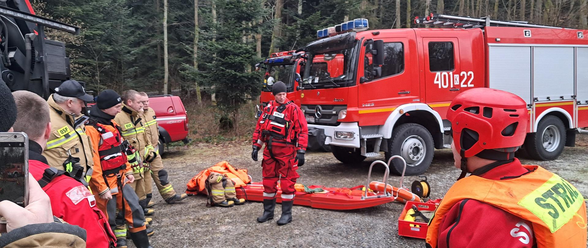 Zdjęcie przedstawia przeprowadzany instruktarz podczas ćwiczeń z zakresu ratownictwa lodowego. Na zdjęciu widać strażaka ubranego w czerwony strój do pracy w wodzie, dodatkowo ma ubraną czerwoną kamizelkę i czapkę na głowie. Po prawej stronie widać odwróconego tyłem strażaka ubranego w czerwone ubranie do pracy w wodzie, na głowie ma kask i dodatkowo ubraną pomarańczową kamizelkę ratunkową z napisem STRAŻ. Na ziemi rozłożony jest sprzęt do ratownictwa wodno-lodowego oraz sanie lodowe. Po lewej stronie widać strażaków ubranych w piaskowe mundury. Za strażakami widać czerwone pojazdy straży pożarnej. 