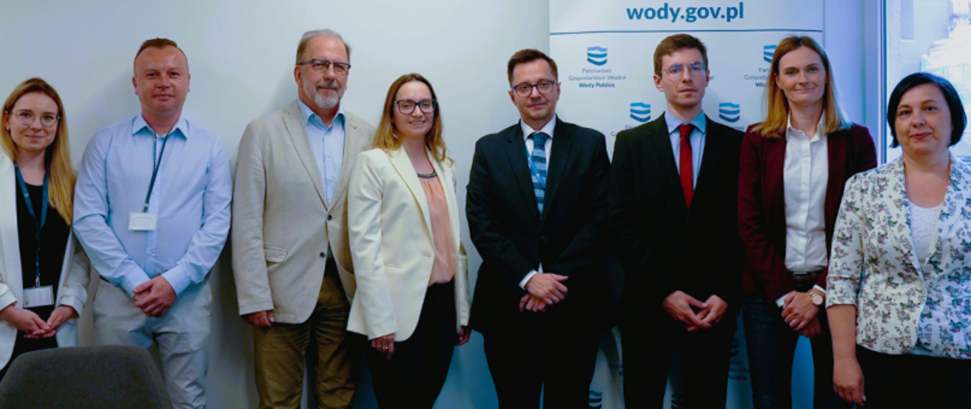 Grupa ośmiu osób w formalnych strojach pozuje do zdjęcia na tle banera „wody.gov.pl” i logo Wód Polskich.