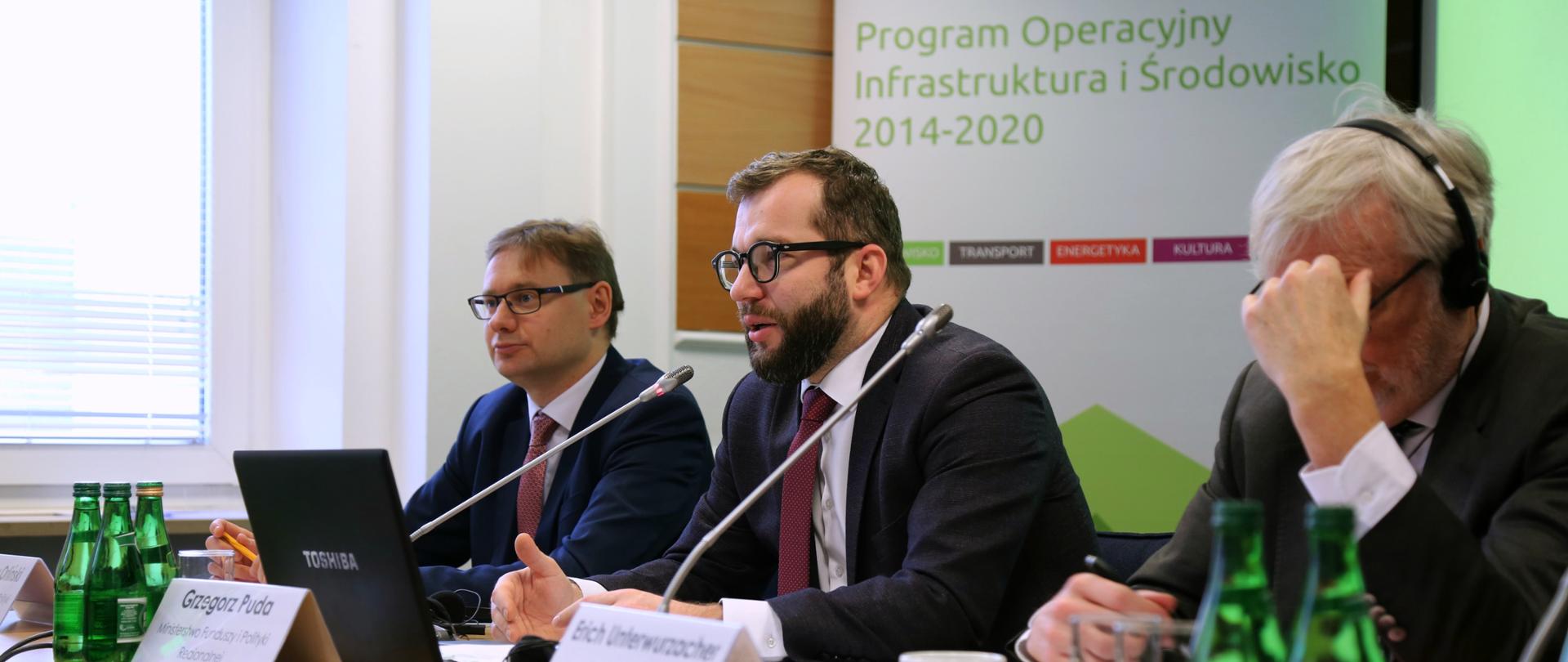 Minister Pudy, dyrektor Orliński i komisarz Underwurzacher siedzą przy stole, za nimi znajduje się napis "Program Infrastruktura i Środowisko" 