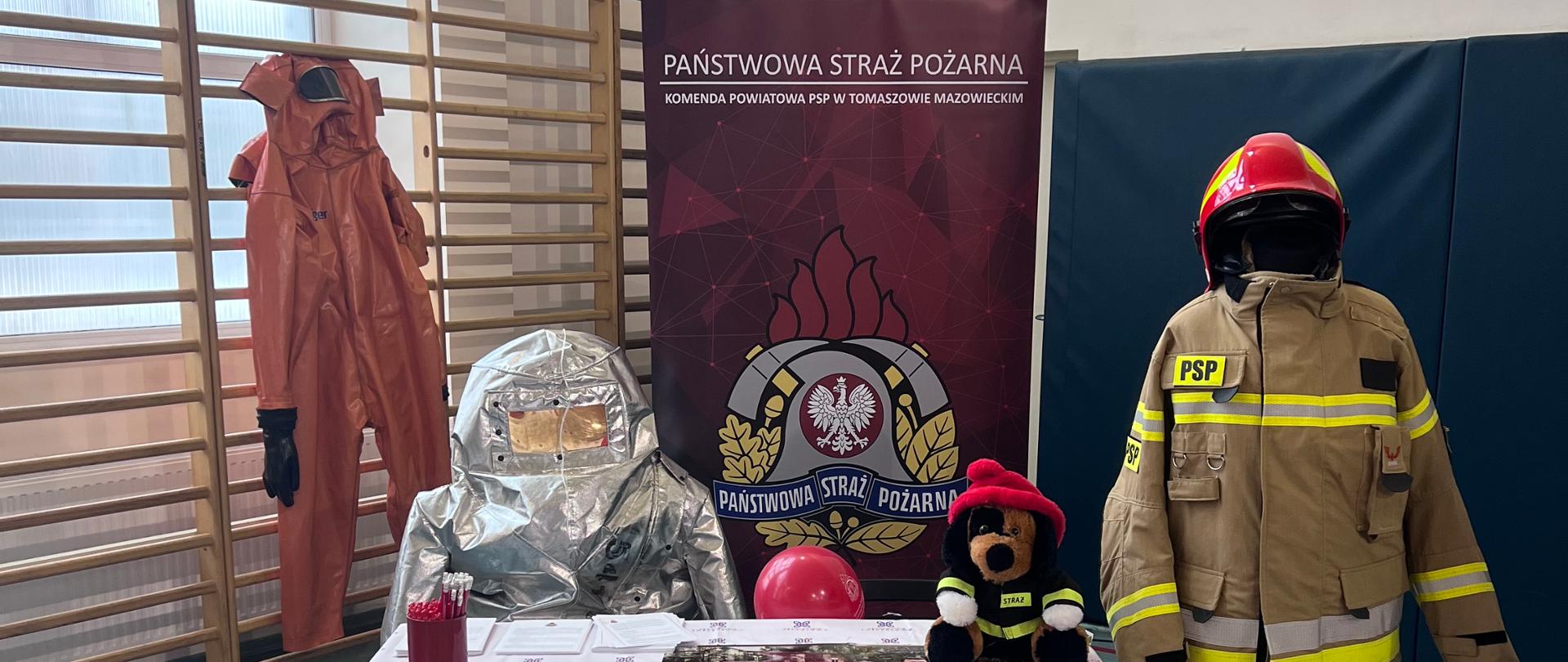 Zdjęcie przedstawia stoisko wystawowe Państwowej Straży Pożarnej z pełnym wyposażeniem ochronnym. Widoczne są manekiny ubrane w specjalistyczne kombinezony oraz strój bojowy strażaka. Na stole znajdują się materiały informacyjne, kalendarz oraz gadżety promocyjne. Całość zaaranżowana jest w przestrzeni sali gimnastycznej.