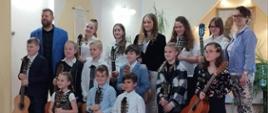 Grupa uczniów trzymających gitary wraz z nauczycielami A. i W. Kozakowskimi uczestników koncertu dla Taty i Mamy na gitarach gramy 26 maja 2023