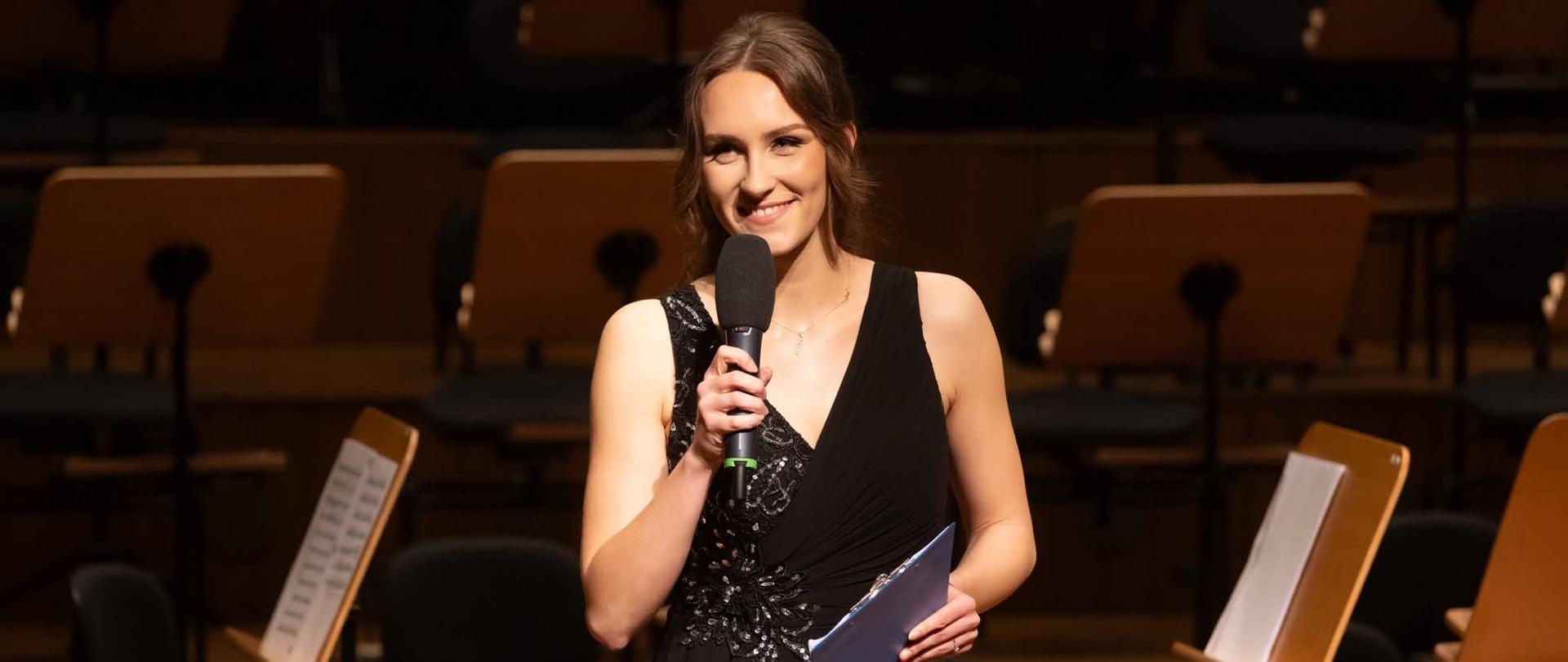 Nauczycielka trzyma mikrofon w ręku, w tle pulpity nut.