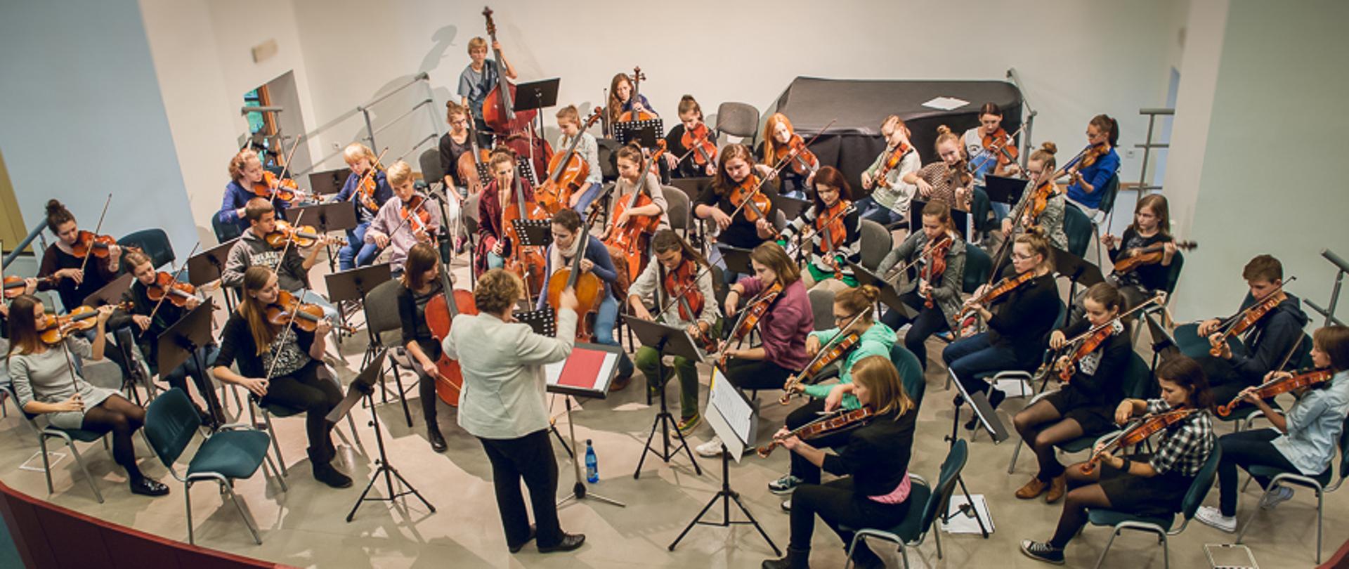 Zdjęcie przedstawia uczniów grających na skrzypcach, altówce, wiolonczeli i kontrabasie oraz dyrygenta podczas próby orkiestry smyczkowej szkoły muzycznej II stopnia w sali koncertowej (zdjęcie kolorowe)