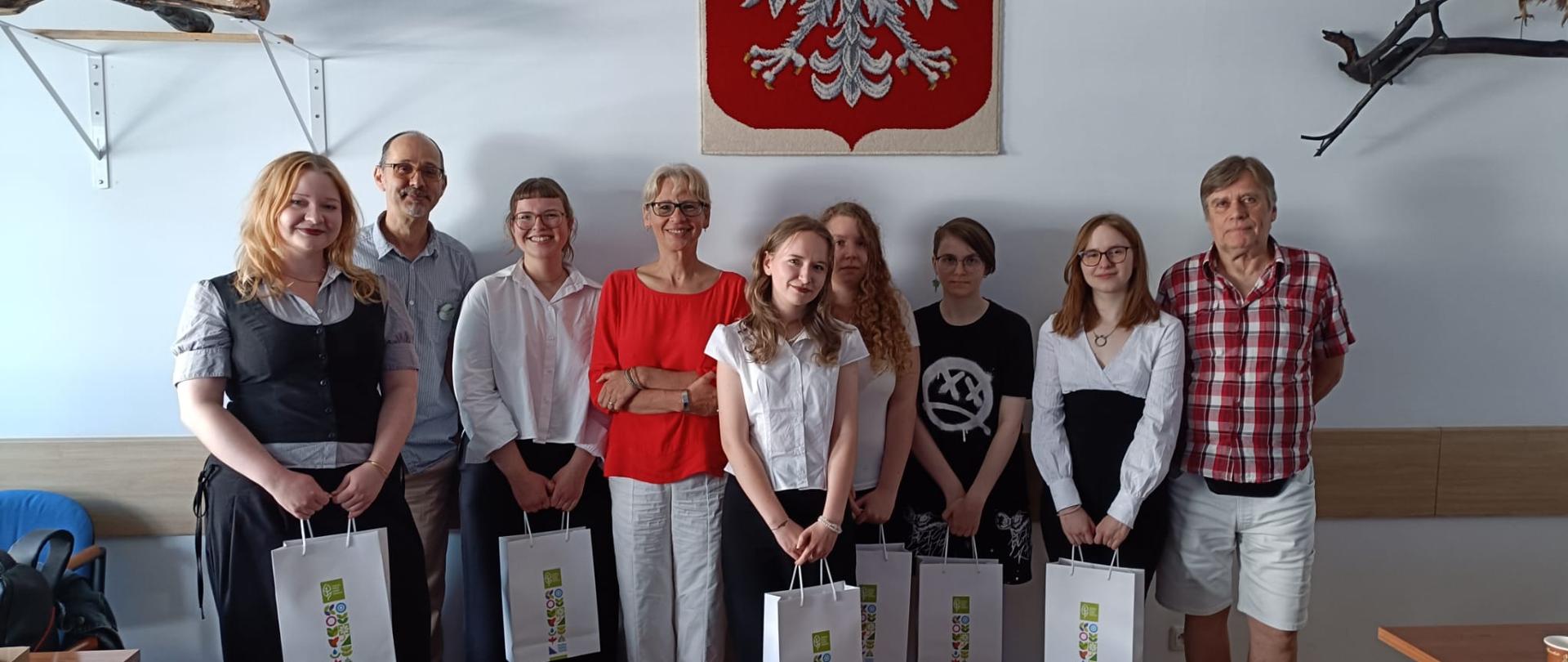 Zdjęcie przedstawia grupę dziewięciu osób stojących w rzędzie w jasnym pomieszczeniu, prawdopodobnie podczas oficjalnego spotkania lub uroczystości. Wśród zebranych znajduje się siedem młodych kobiet oraz dwóch starszych mężczyzn. Część dziewcząt ubrana jest w eleganckie, galowe stroje – białe bluzki oraz ciemne spódnice lub spodnie – i trzyma w dłoniach jednakowe, białe torby upominkowe z kolorowym nadrukiem. Pozostałe osoby noszą bardziej casualowe ubrania, takie jak czerwona bluzka, koszula w kratę czy czarny t shirt z grafiką. W centralnym punkcie białej ściany za nimi wisi godło Polski przedstawiające białego orła w koronie na czerwonej tarczy, a po bokach widoczne są elementy dekoracyjne z drewna i gałęzi. W tle po lewej i prawej stronie kadru można dostrzec fragmenty biurek oraz krzeseł, co sugeruje, że spotkanie odbywa się w klasie lub sali konferencyjnej.
