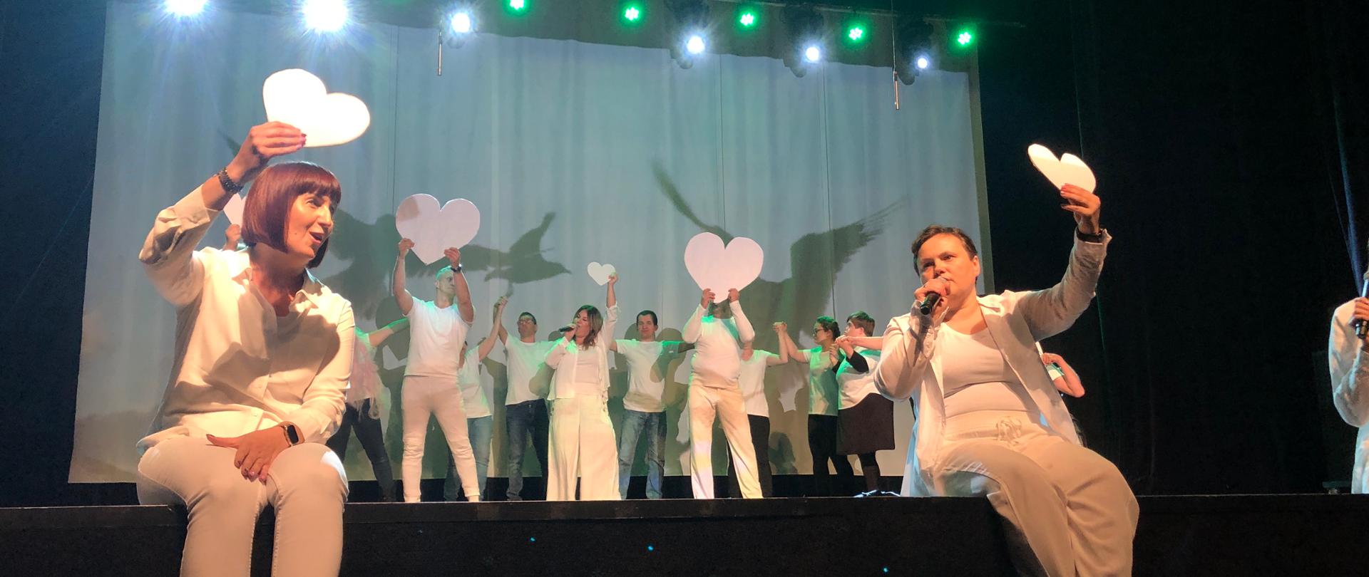 Aktorzy na scenie. W dłoniach trzymają białe papierowe serca. 