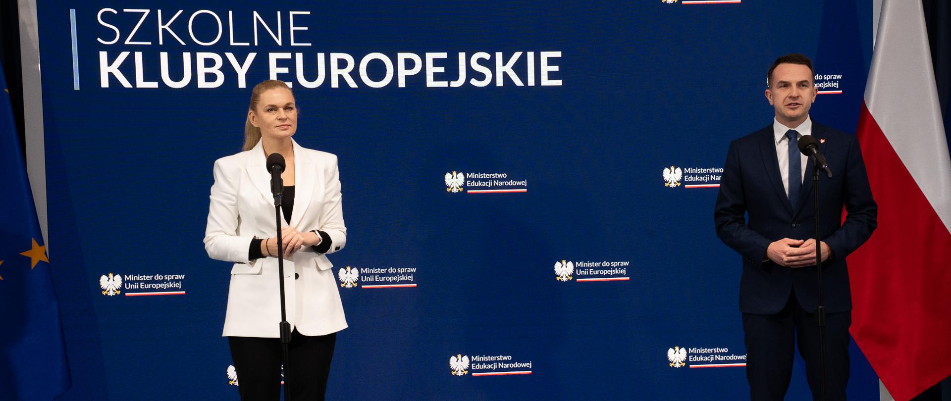 Na tle wbudowanego w ścianę wielkiego ekranu z napisem Szkolne Kluby Europejskie stoi minister Nowacka, obok niej minister Adam Szłapka. Oboje mają przed sobą ustawione mikrofony na stojaku.