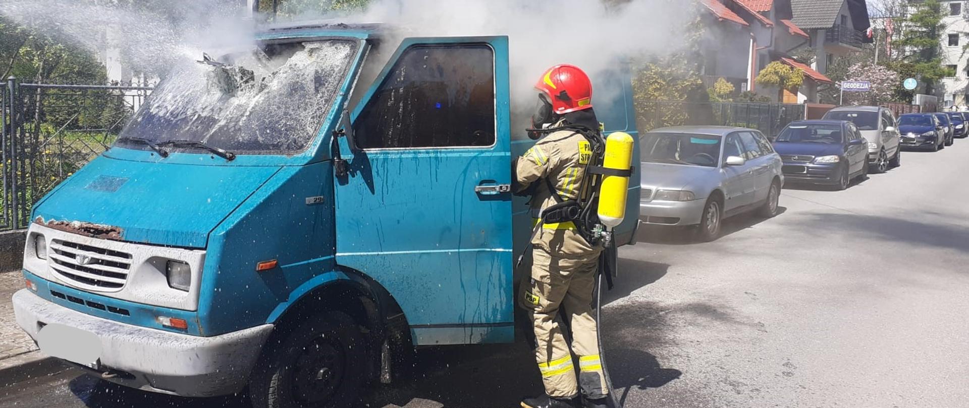 Na zdjęciu widzimy samochód dostawczy w kolorze zielonym z którego wydobywa się dym. Z boku samochodu stoi strażak w ubraniu ochronnym i aparacie ochrony dróg oddechowych prowadzący działania gaśnicze w kabinie. W tle widoczne są zabudowania ulicy oraz zaparkowane pojazdy.